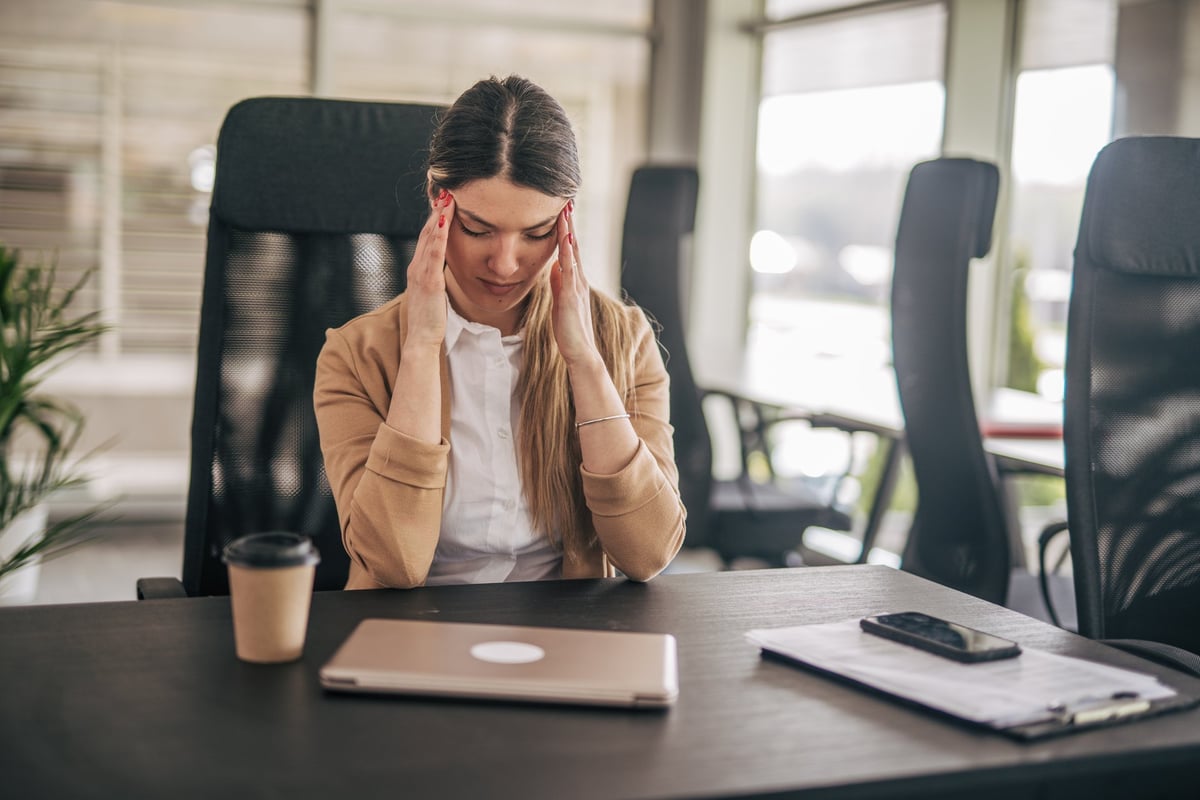 Young businesswoman sitting at her desk with a laptop, documents and smartphone, suffering from a headache and touching her temples with her hands