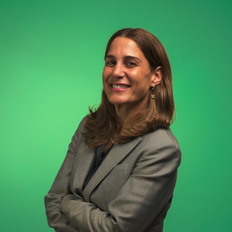 Woman with brown shoulder-length hair smiling and standing with arms crossed against a green background, wearing a gray blazer