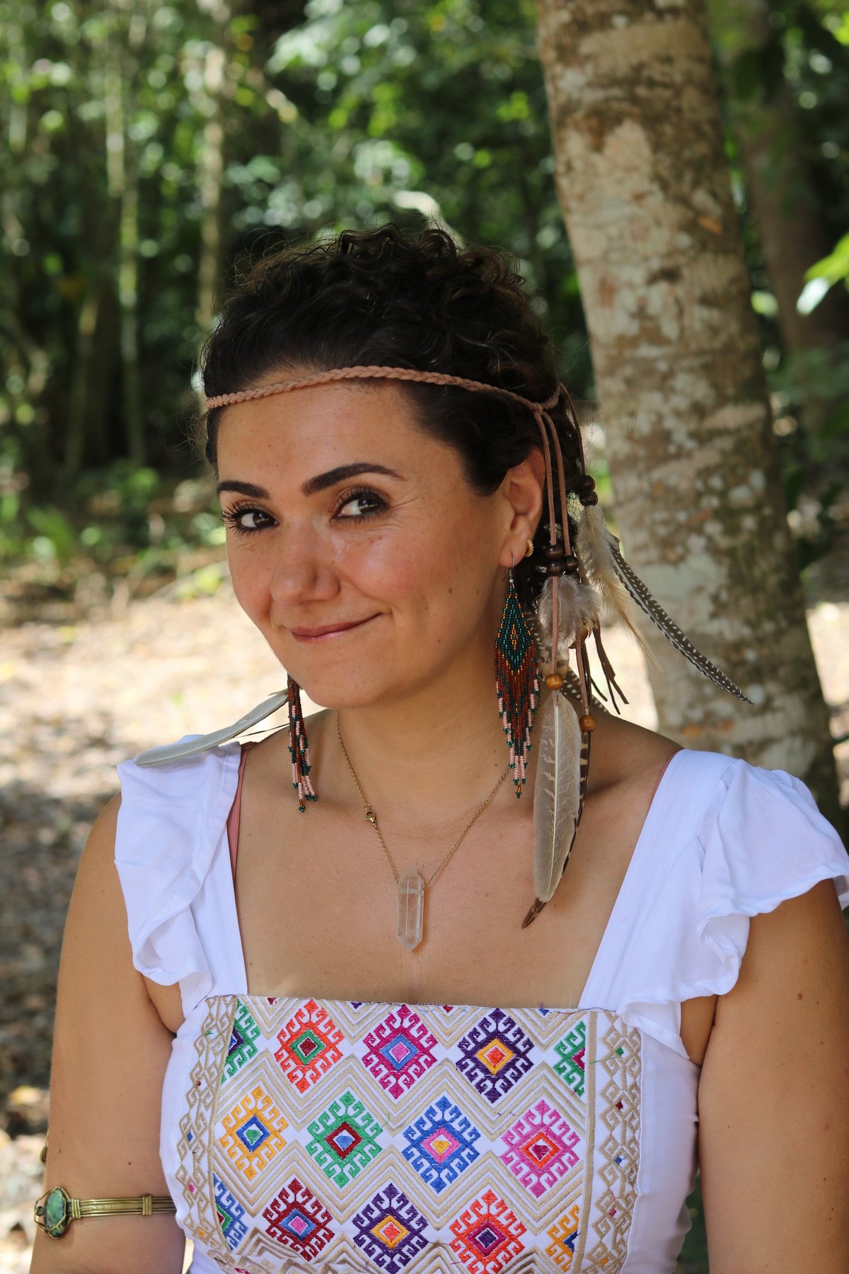 Woman smiling outdoors wearing white top with colorful geometric embroidery, braided hair with beaded decorations, forest background
